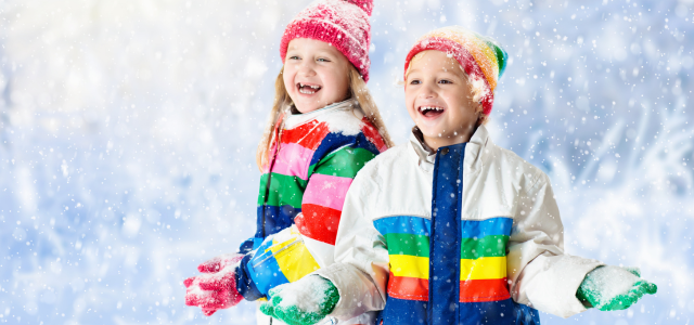 Zwei Kinder in bunten Winterjacken und Mützen lachen im fallenden Schnee und strecken ihre Hände nach den Schneeflocken aus.