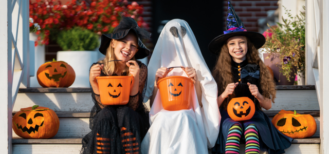 Drei verkleidete Kinder sitzen auf einer Treppe mit Halloween-Kürbissen, lachen und halten orangefarbene Süßigkeiteneimer, darunter ein Kind als Geist und zwei als Hexen.