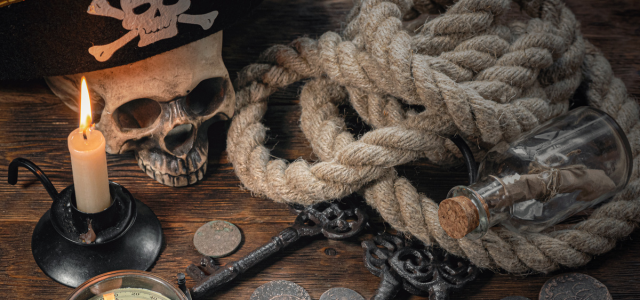 Stillleben mit Piratenmotiven: Totenschädel mit Hut, brennende Kerze, dickes Tau, alte Schlüssel, Münzen und eine Flaschenpost auf einem Holztisch.